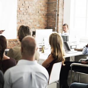 A lecturer speaks at the front of a classroom to a group of engaged and listening professionals. 