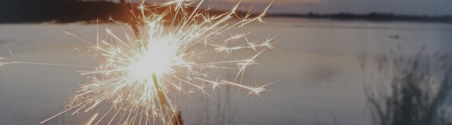 A lit sparkler providing illumination at dusk overlooking water