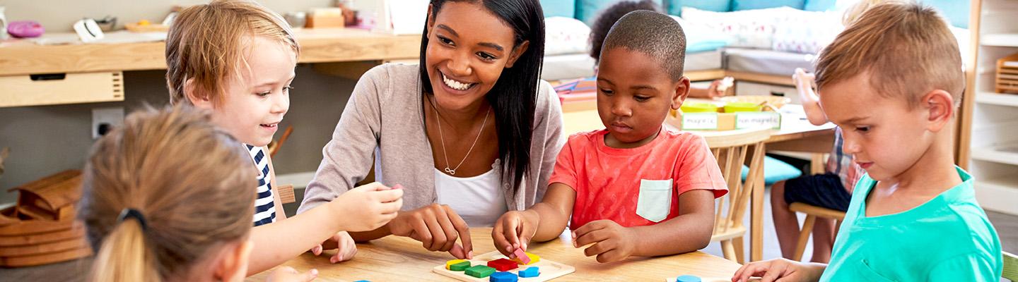 Group of young students gathered in a workgroup with shape and color puzzles, assisted by an encouraging teacher